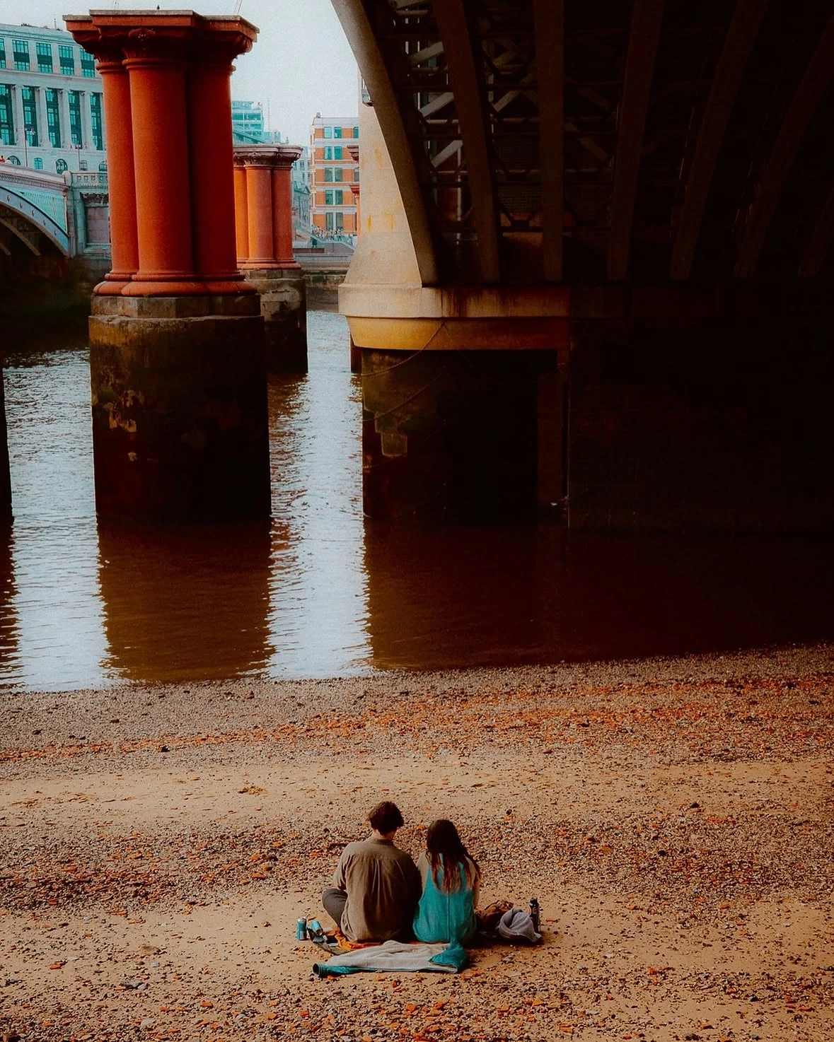 A couple sitting beside the Thames beneath a London bridge.