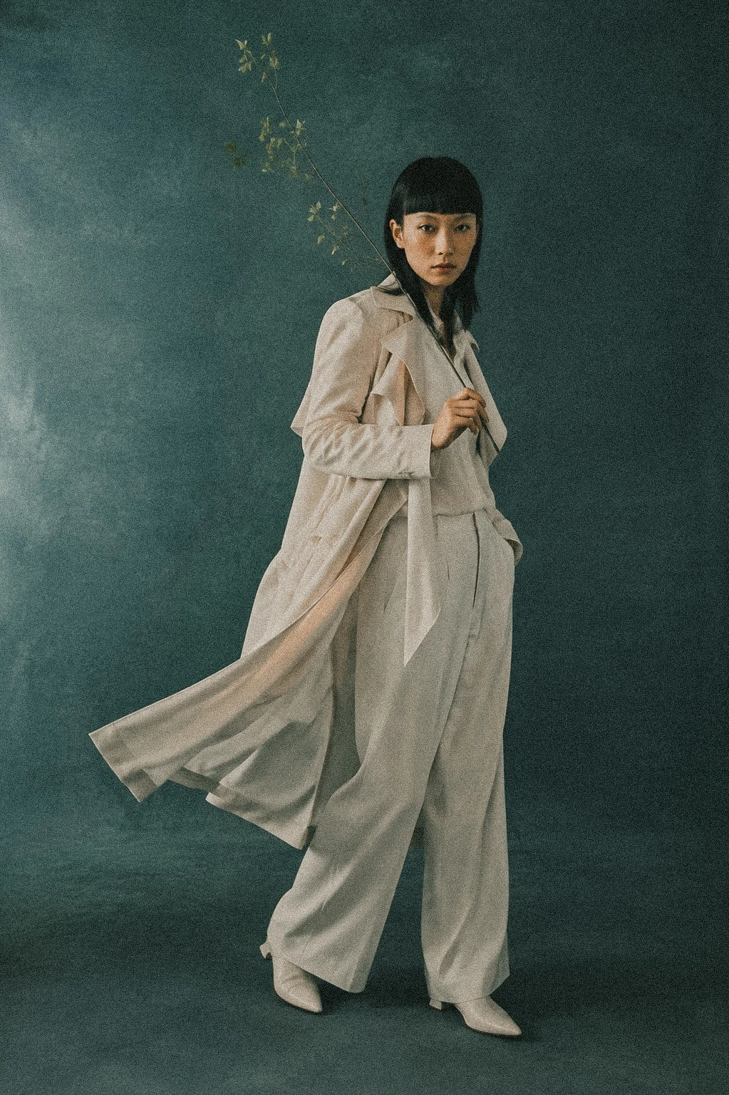 Cream tailoring and a flowing coat against a teal studio backdrop.