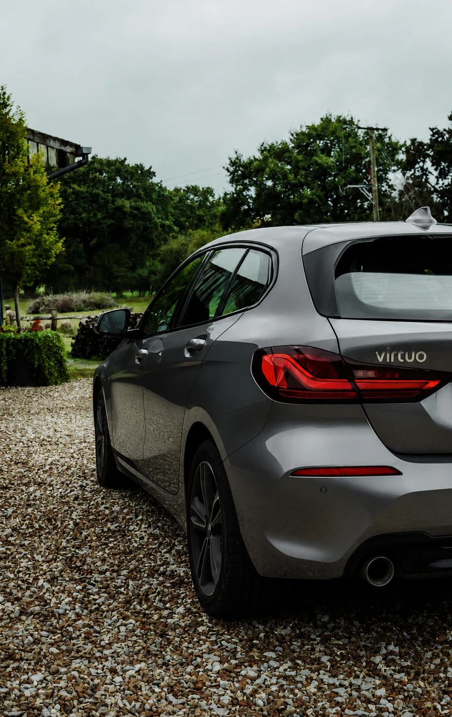 A deep silver Virtuo car parked on gravel beside a garden.